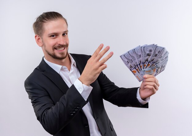 Handsome business man wearing suit holding cash showing and piinting up with fingers number three looking surprised standing over white background