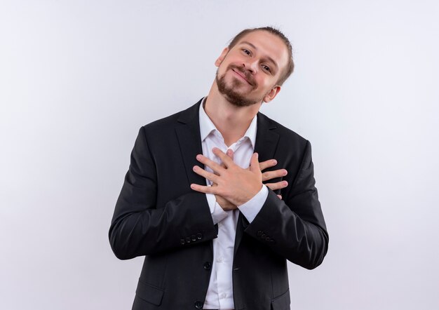 Handsome business man wearing suit holding hands on chest feeling positive emotions standing over white background