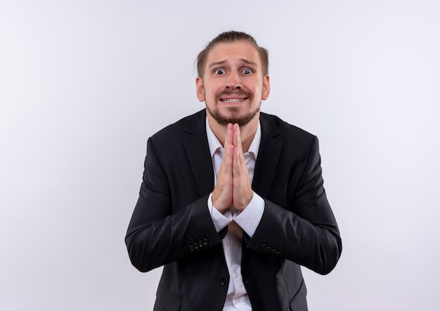 Handsome business man wearing suit holding hands together praying and begging with hope expression standing over white background