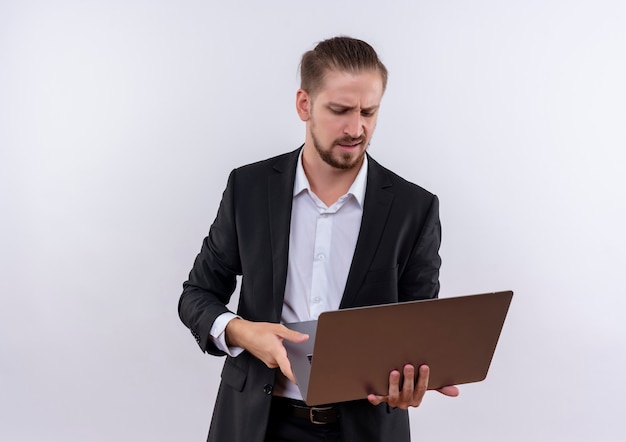 白い背景の上に立って不機嫌な画面を見ているラップトップコンピューターを保持しているスーツを着ているハンサムなビジネスマン