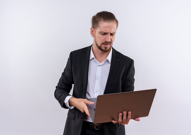 Handsome business man wearing suit holding laptop computer looking at screen displeased standing over white background