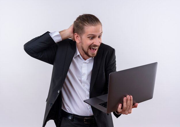 Handsome business man wearing suit holding laptop computer looking surprised and amazed standing over white background