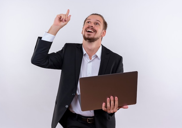 흰색 배경 위에 유쾌하게 서있는 손가락으로 가리키는 노트북 컴퓨터를 들고 양복을 입고 잘 생긴 비즈니스 남자