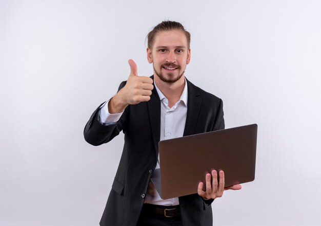 Красивый деловой человек в костюме, держащий портативный компьютер, весело улыбаясь, показывает палец вверх, глядя на камеру, стоящую на белом фоне