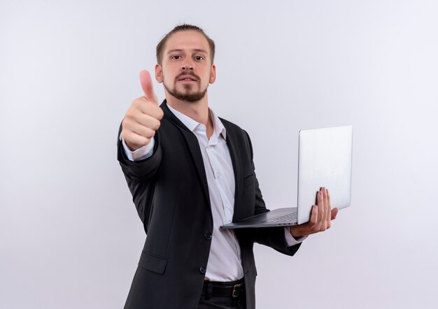 Красивый деловой человек в костюме держит портативный компьютер, весело улыбаясь, показывает палец вверх, стоя на белом фоне