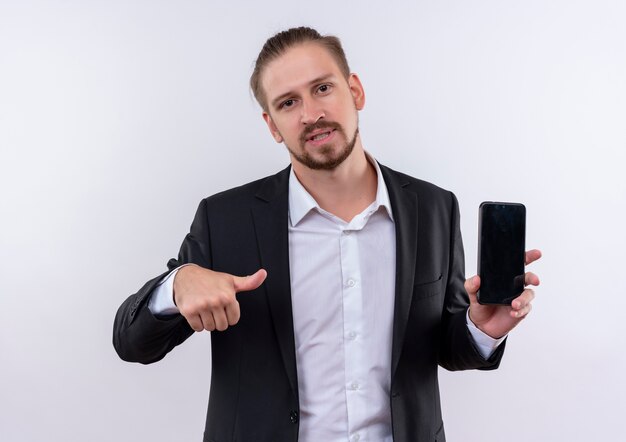 Красивый деловой человек в костюме держит смартфон, указывая пальцем на него, выглядит уверенно, стоя на белом фоне