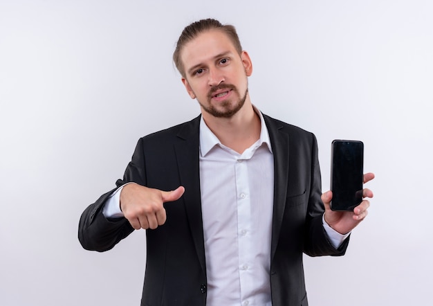 Handsome business man wearing suit holding smartphone pointing with thumb to it looking confident standing over white background