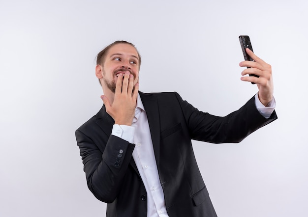 白い背景の上に立っている顔に恥ずかしがり屋の笑顔でselfieを取っているスマートフォンを保持しているスーツを着ているハンサムなビジネスマン