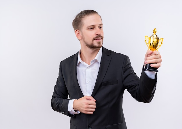 Бесплатное фото Красивый деловой человек в костюме держит трофей, глядя на него с улыбкой на лице, стоя на белом фоне