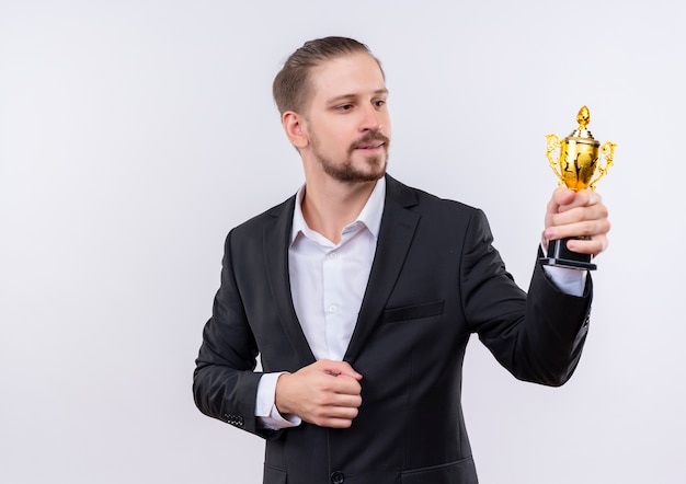 Handsome business man wearing suit holding trophy looking at it with smile on face standing over white background