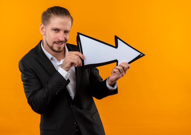 Handsome business man wearing suit holding white arrow looking at camera smiling and winking standing over orange background