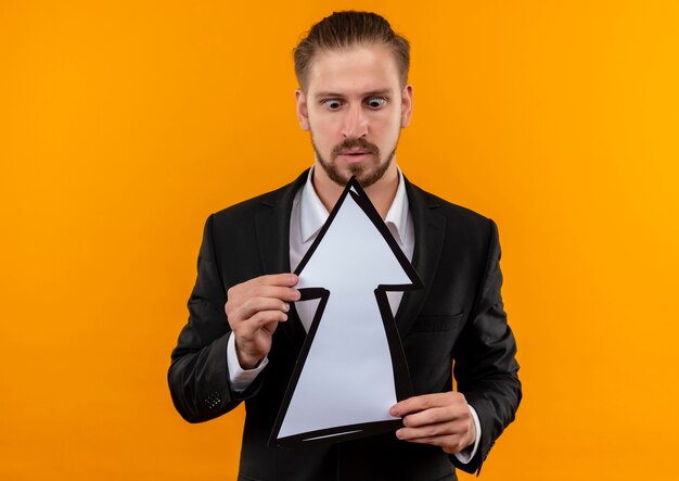 Handsome business man wearing suit holding white arrow looking at it confused and surprised standing over orange background
