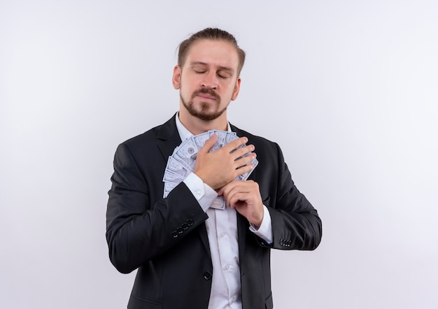 Handsome business man wearing suit hugging cash holding money on his chest with closed eyes standing over white background