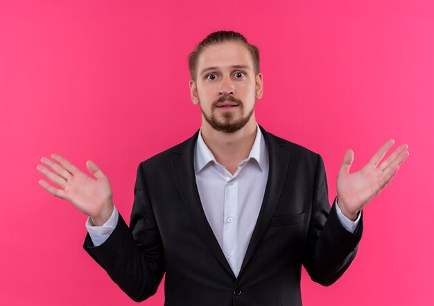 Handsome business man wearing suit loking at camera confused with raised hands standing over pink background