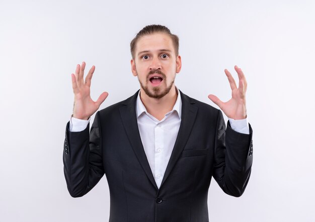 Handsome business man wearing suit looking at camera disappointed raising arms standing over white background