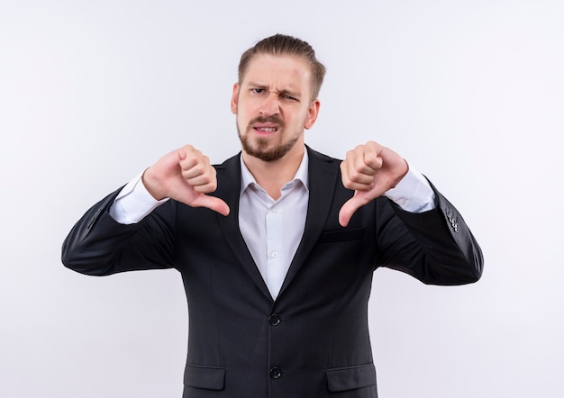 Handsome business man wearing suit looking at camera displeased showing thumbs down standing over white background