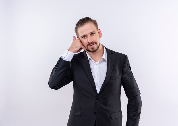 Handsome business man wearing suit looking at camera smiling making call me gesture standing over white background