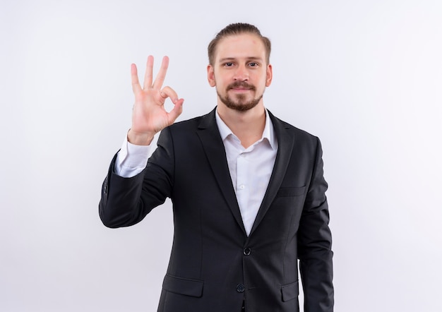 Handsome business man wearing suit looking at camera smiling showing ok sing standing over white background