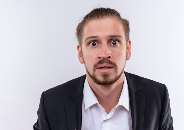 Handsome business man wearing suit looking at camera surprised and amazed standing over white background