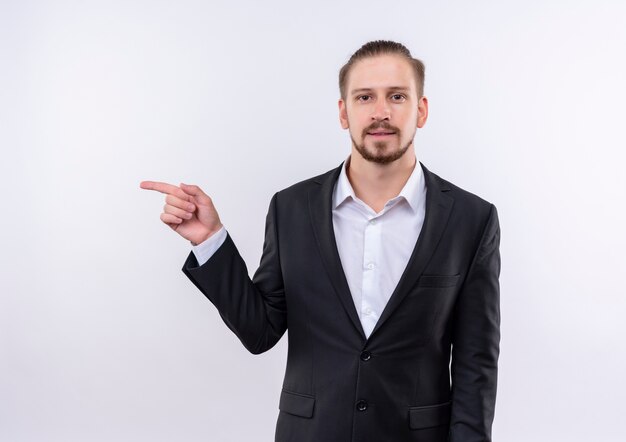 Handsome business man wearing suit looking at camera with smile on face pointing with finger to the side standing over white background