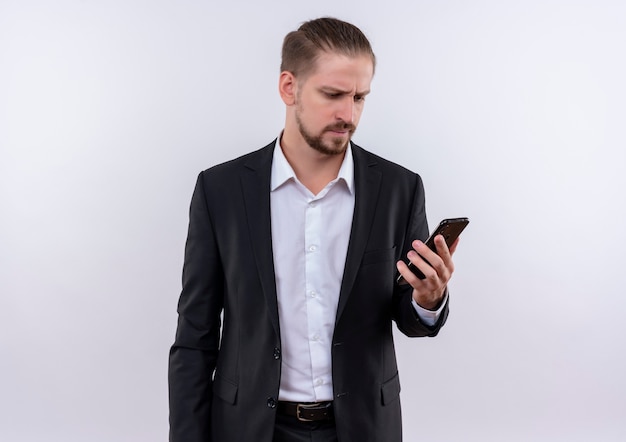 Handsome business man wearing suit looking at his smartphone confused standing over white background