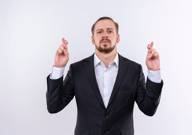 Handsome business man wearing suit making desirable wish crossing fingers standing over white background