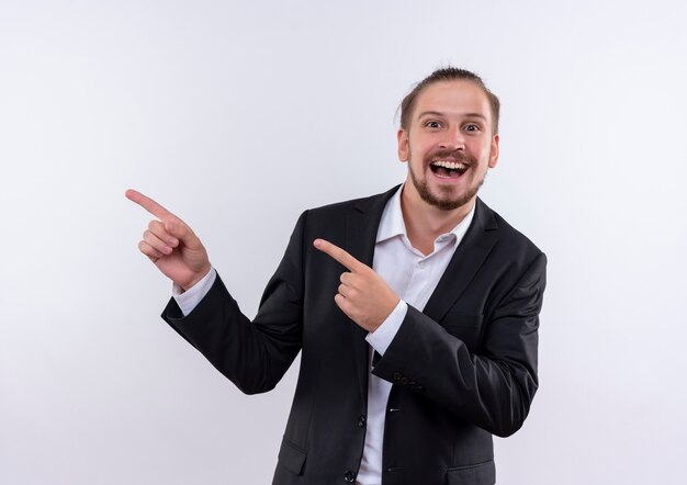 Handsome business man wearing suit pointing with fingers to the side looking at camera with big smile on face standing over white background