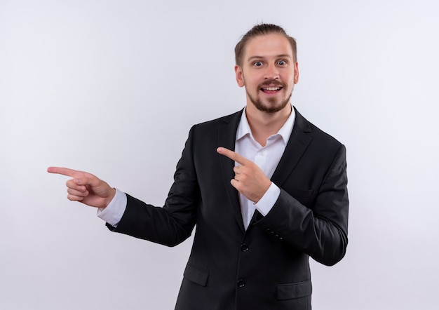 Handsome business man wearing suit pointing with fingers to the side looking at camera with smile on face standing over white background