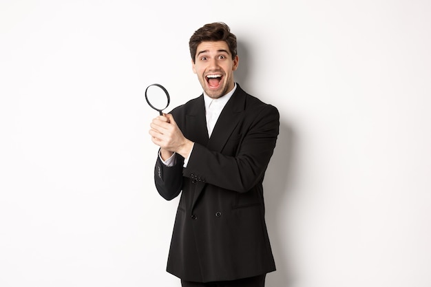 Handsome businessman in black suit, holding magnifying glass and smiling, found something, standing against white background