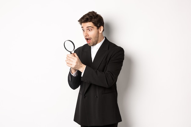 Free photo handsome businessman in black suit, holding magnifying glass and smiling, found something, standing against white background.