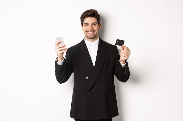 Handsome businessman in black suit smiling, showing credit card and money, standing against white background