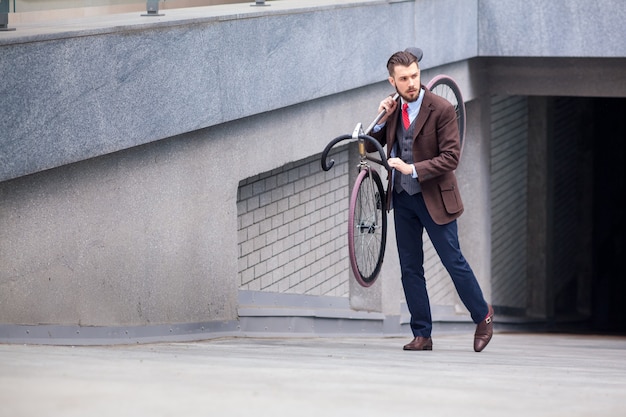 街の通りで彼の自転車を運ぶハンサムな実業家。若い男性の現代的なライフスタイルの概念