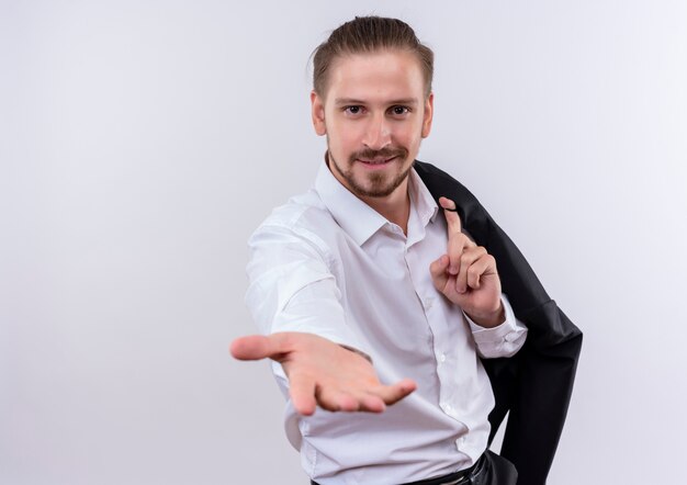 Handsome businessman carrying his jacket on shoulder looking at camera smiling friendly offering hand standing over white background