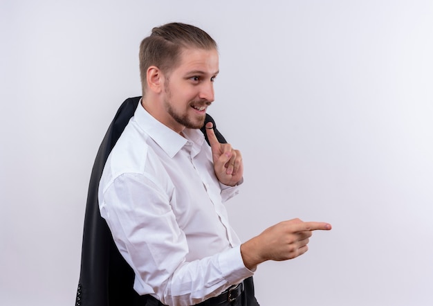 Handsome businessman carrying his jacket on shoulder smiling looking aside pointing with finger to the side standing over white background