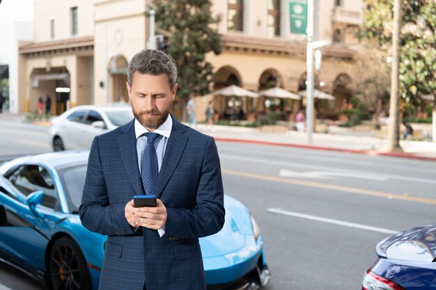 스마트폰에서 채팅을 하는 잘생긴 사업가는 야외 고급 자동차 옆에 서 있습니다. 차량 이용 가능. 근처에 비싼 양복을 입은 남자. 자동차 보험 사업. 성공한 딜러. 자신감 있는 매니저 렌탈 온라인