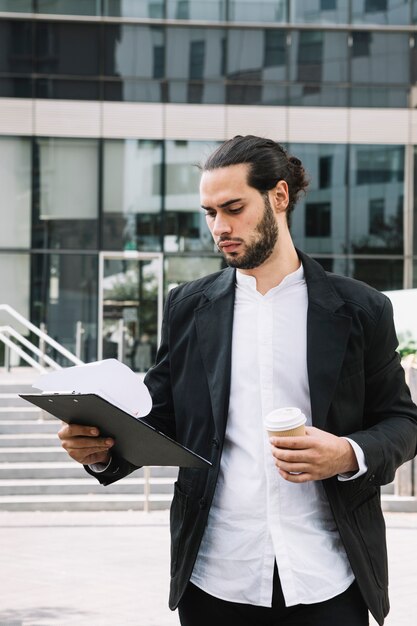 Handsome businessman holding takeaway coffee cup looking at clipboard