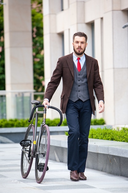 Красивый бизнесмен в куртке и красном галстуке и его велосипеде на улицах города.