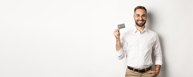 Handsome businessman showing his credit card looking satisfied standing over white background