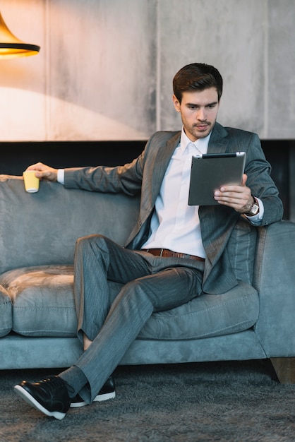 デジタルタブレットを見る使い捨てカップを持っているソファに座っている幸せなビジネスマン