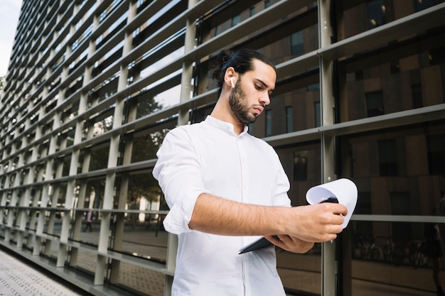 Handsome businessman standing outside the corporate building looking at documents on clipboard