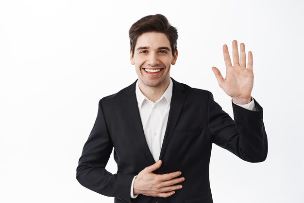 Handsome businessman in suit, corporate man waving hand and say hello, greet you, introduce himself on party, wearing formal clothes, standing over white background