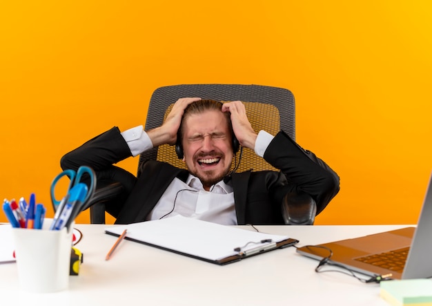 Handsome businessman in suit and headphones with a microphone going wild by pulling his hair sitting at the table in offise over orange background