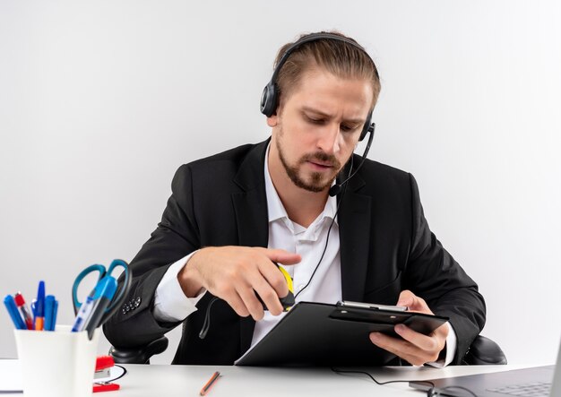 Handsome businessman in suit and headphones with a microphone holding clipboard looking at it with serious face sitting at the table in offise over white background