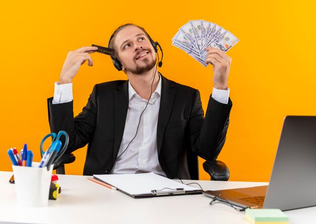 Handsome businessman in suit and headphones with a microphone holding cush and credit card with dreamy look sitting at the table in offise over orange background