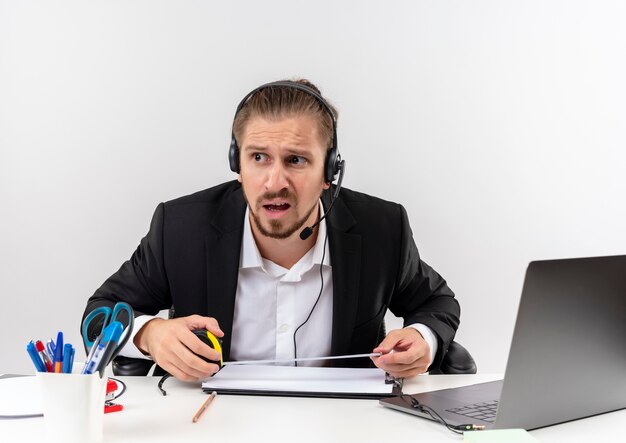 Handsome businessman in suit and headphones with a microphone looking aside confused sitting at the table in offise over white background