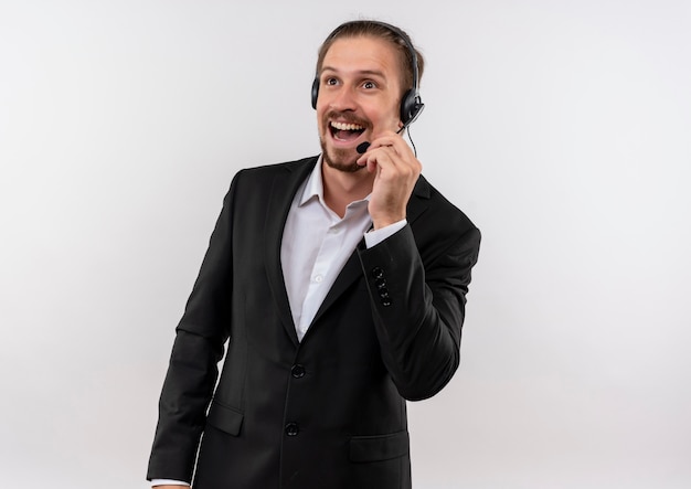 Handsome businessman in suit and headphones with a microphone looking aside listening to a client smiling with happy face standing over white background