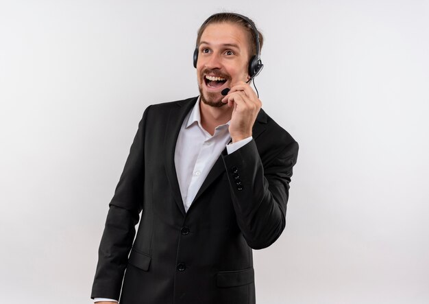 Handsome businessman in suit and headphones with a microphone looking aside listening to a client smiling with happy face standing over white background