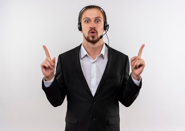 Handsome businessman in suit and headphones with a microphone looking at camera pointing up with index fingers surprised and happy standing over white background