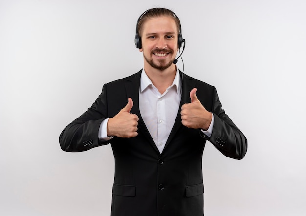 Handsome businessman in suit and headphones with a microphone looking at camera smiling showing thumbs up standing over white background