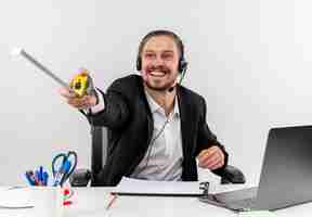 Free photo handsome businessman in suit and headphones with a microphone pointing with measure tape to the side with smile on face sitting at the table in offise over white background